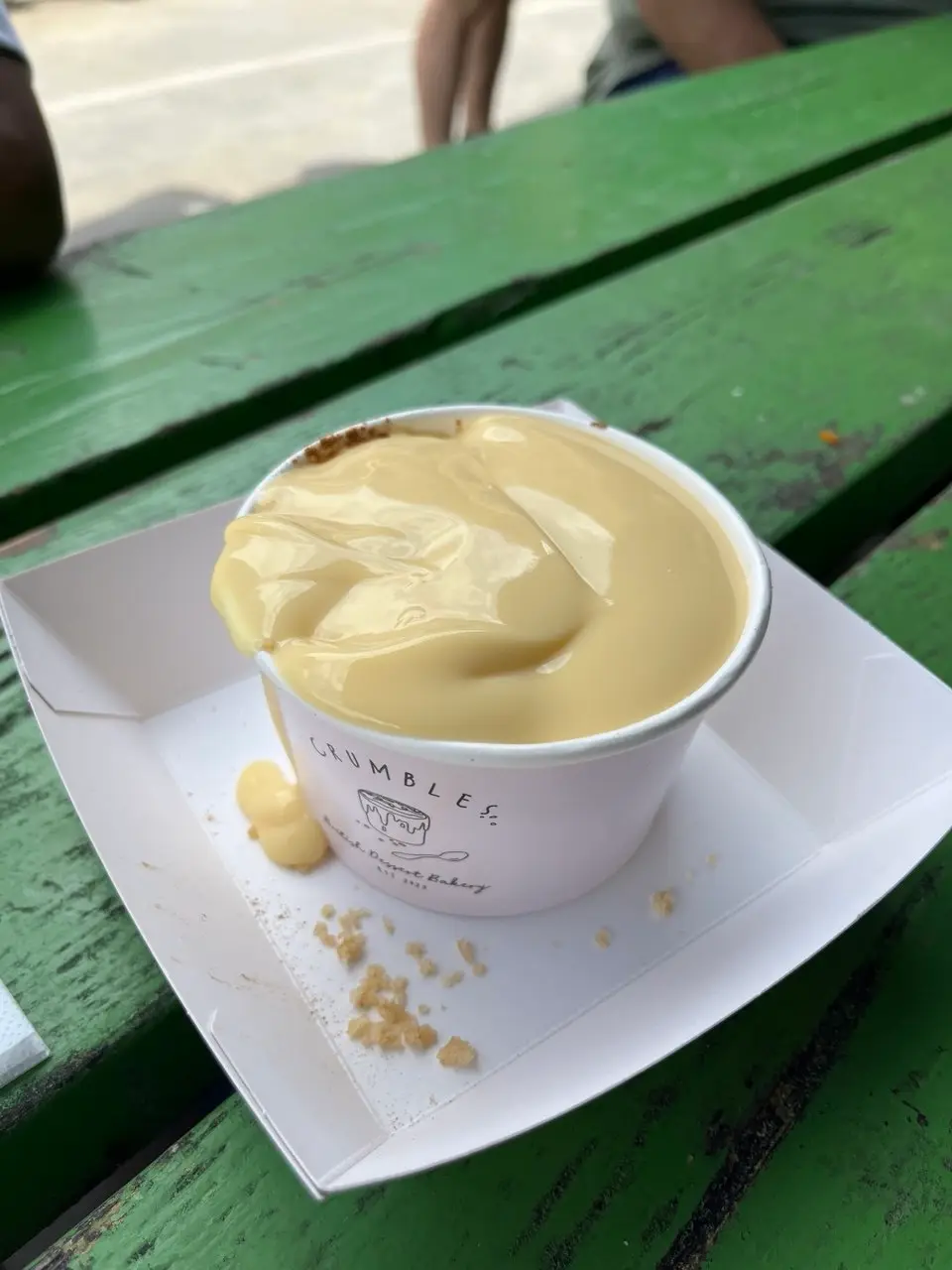 A paper tub of apple crumble under a thick pour of custard, on a tray on a green picnic bench