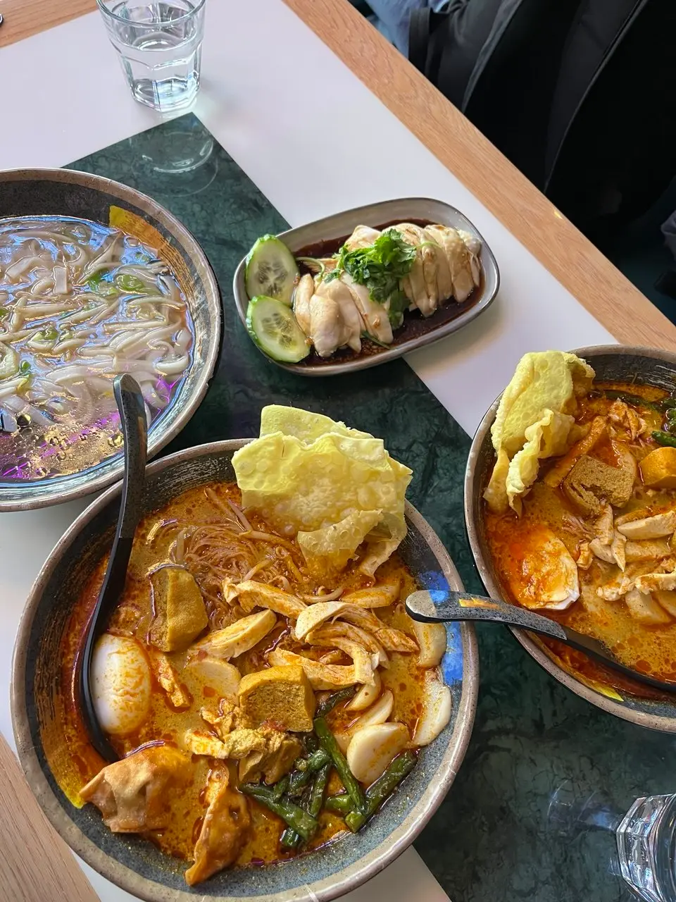 Two bowls of mee kari in dark stoneware, orange coconut broth with shredded chicken, tofu puffs, fish cake and long beans, a sheet of crisp bean curd propped on top of each
